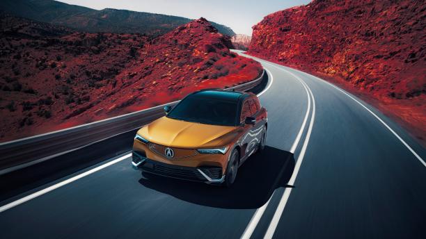 3/4 bird’s eye front view of gold ZDX driving on mountain highway. Part of the image is saturated with colour, so the dirt on the sides of the highway is red.