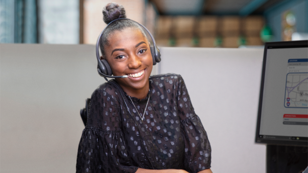 A young woman sat at a desk in an office, with telephone headset on, smiles at camera.