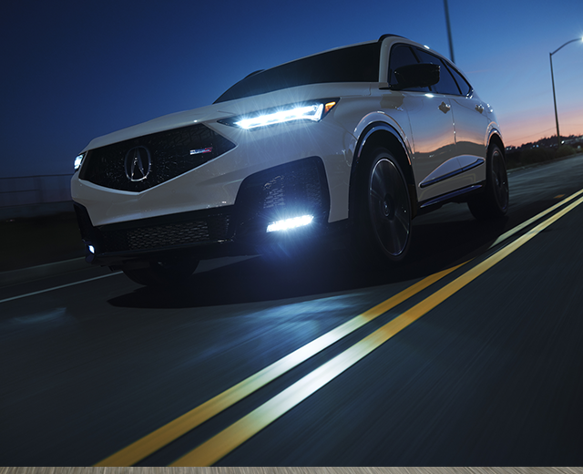 A white 2025 MDX Type S drives on an illuminated city highway at night with headlights on.