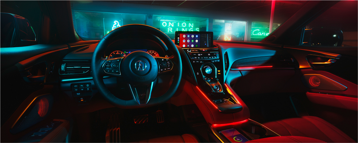 Plan panoramique intérieur du volant et du tableau de bord. À travers le pare-brise, on voit que le RDX est stationné devant un restaurant éclairé par des néons, la nuit.