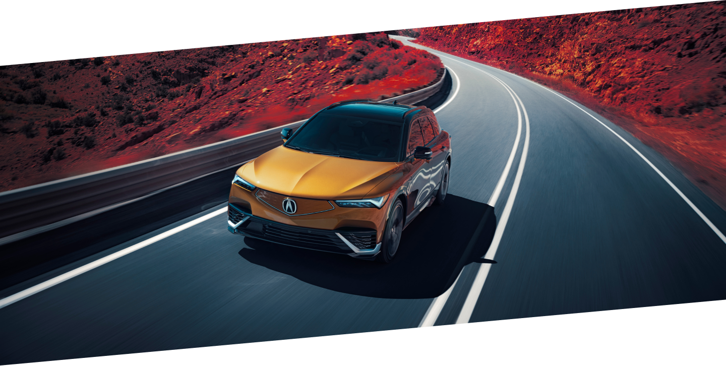 3/4 bird’s eye front view of gold ZDX driving on mountain highway. Part of the image is saturated with colour, so the dirt on the sides of the highway is red.