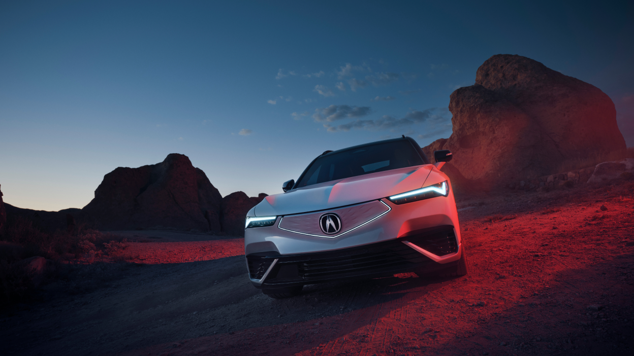 Medium wide front view of white ZDX parked in the desert at dusk. Its headlights are on and the diamond pentagon front fascia is illuminated.