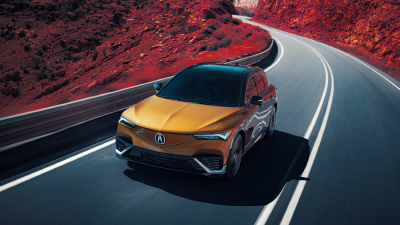 3/4 bird’s eye front view of gold ZDX driving on mountain highway. Part of the image is saturated with colour, so the dirt on the sides of the highway is red.