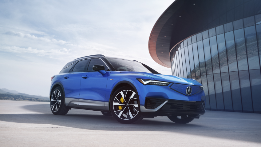 3/4 front view of a blue ZDX parked outside a modern glass-walled building at sunset.