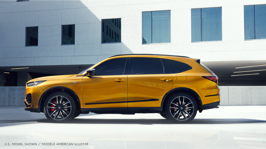 Plan étendu latéral d’un MDX Type S de couleur or à l’arrêt dans un stationnement situé à l’extérieur d’un bâtiment moderne de couleur blanche au style minimaliste.