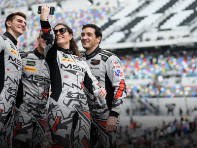Group shot of an Acura pit crew taking a selfie on the track with stadium seating visible in the background. Photo d’une équipe de ravitaillement Acura prenant un égoportrait sur un circuit de course avec des estrades en arrière-plan.
